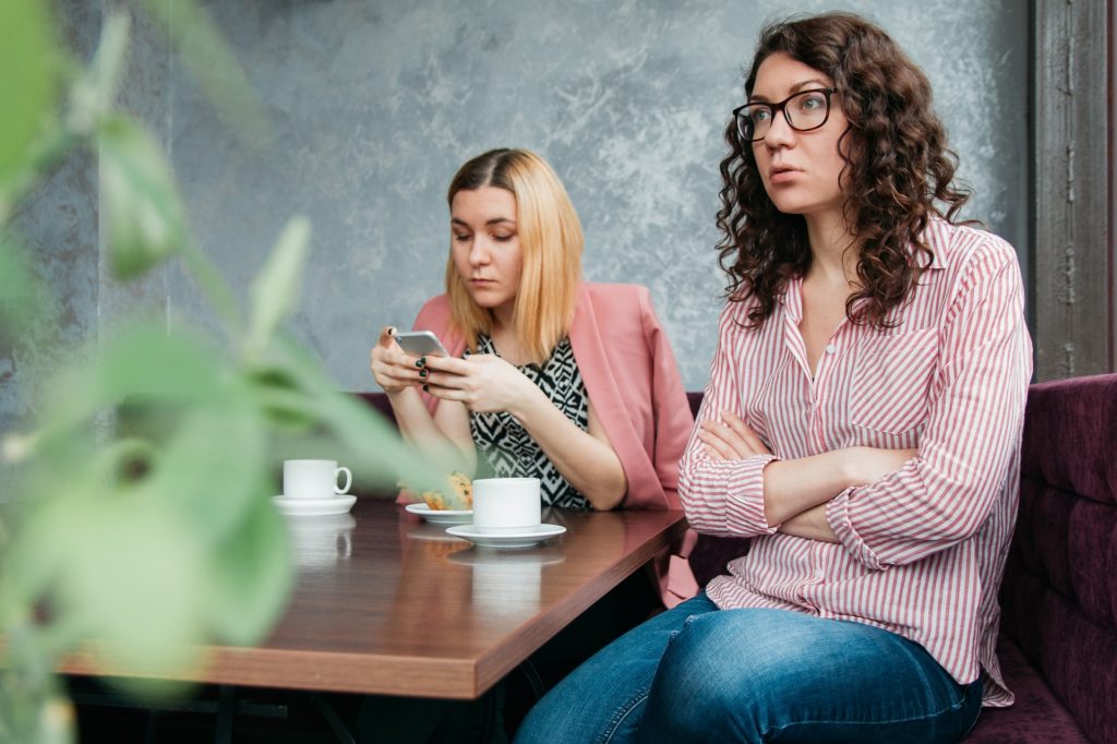 To kvinner på kafé, der den ene sitter opptatt av telefonen, og den andre virker veldig lite fornøyd med det.